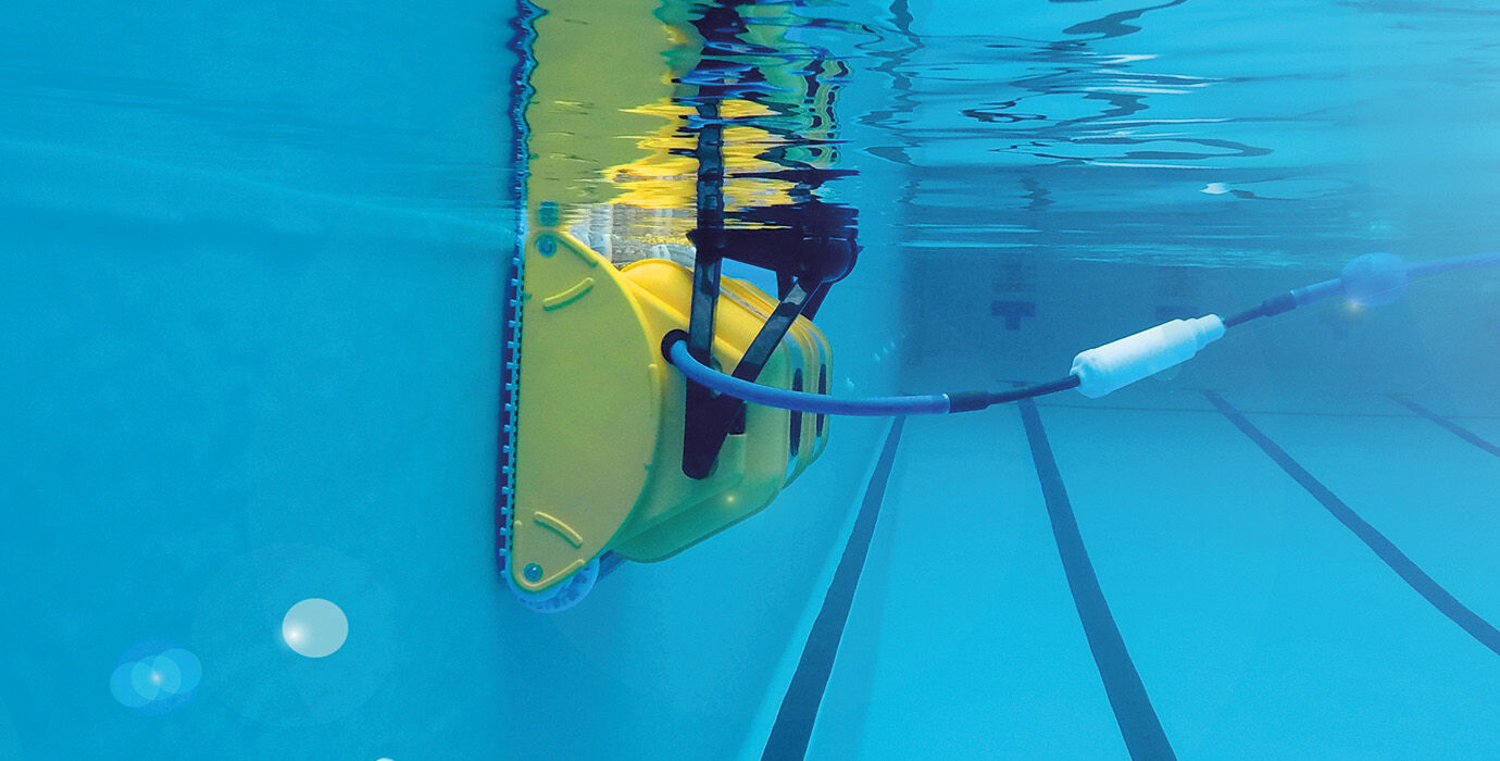 Yellow robotic pool cleaner with a black and white hose attachment, mounted against the tiled wall of an underwater swimming pool.