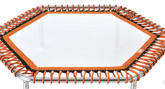 Hexagonal trampoline with orange elastic edges and a white jumping surface, framed by silver metal poles.